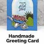 Handmade card in blues, greens and pinks featuring a wheelbarrow with flowers underneath an arch with sentiment "every day is a fresh start."